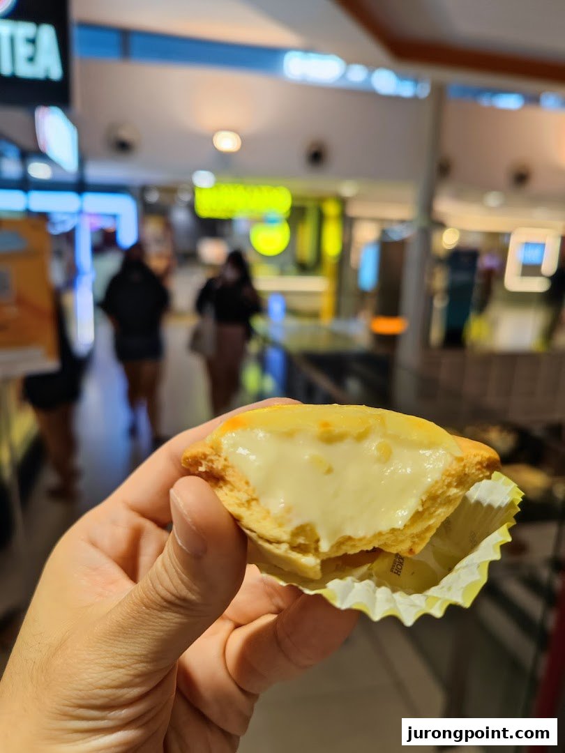 A hand holds a delicious Hokkaido Baked Cheese Tart, showcasing its creamy filling in a mall setting.
