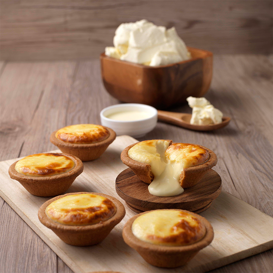 Several golden-baked cheese tarts, one cut open revealing a creamy, molten cheese filling, with ingredients in the background.