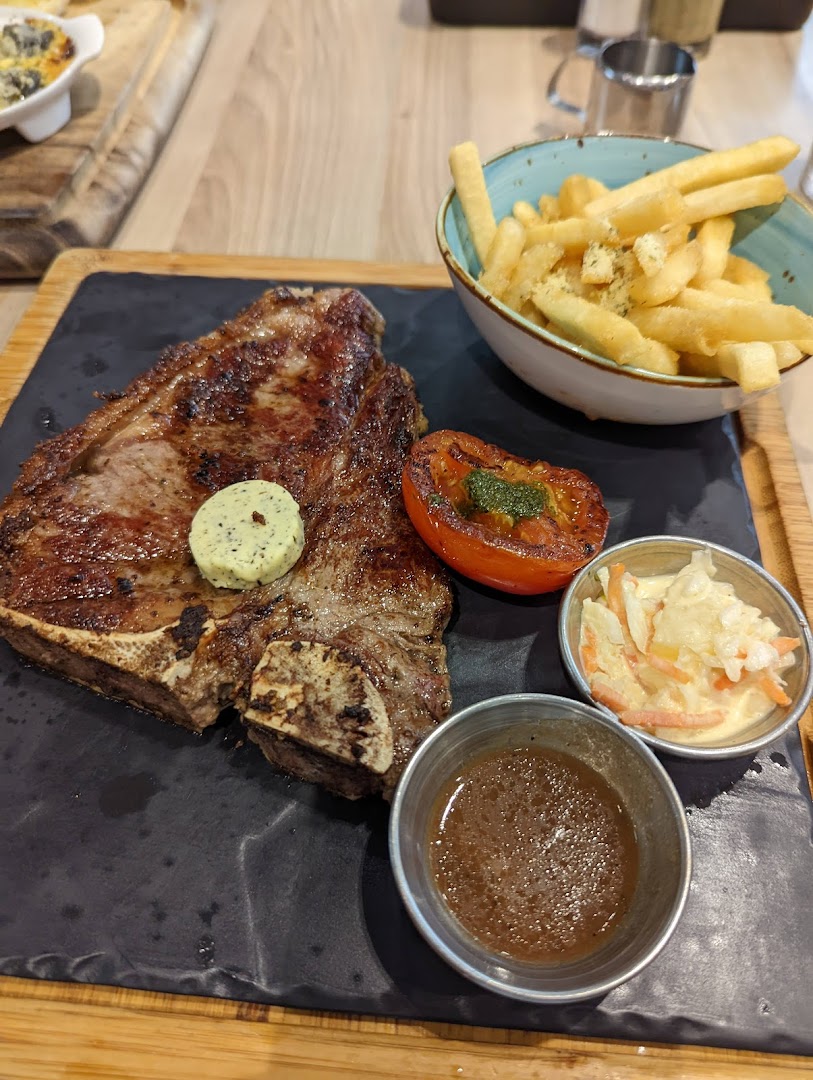 A perfectly grilled T-bone steak served with golden fries, roasted tomato, coleslaw, and sauce.