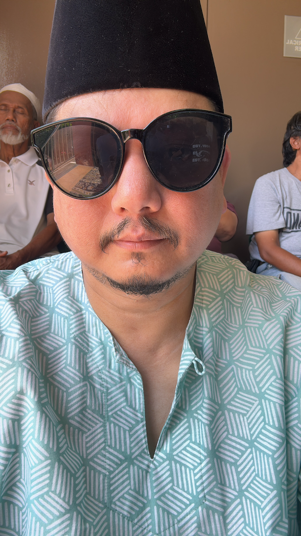 A person wearing sunglasses and a traditional hat, with other individuals in the background.