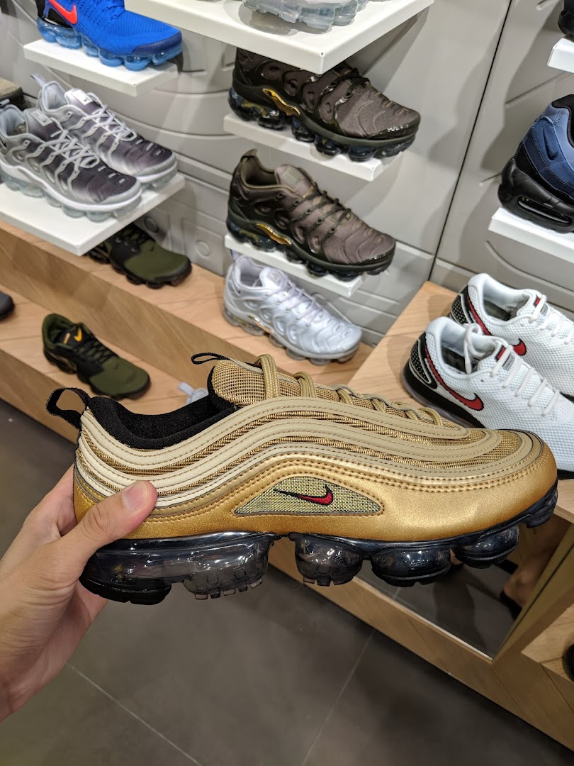 A hand holds a gold Nike sneaker in a JD Sports store, surrounded by other athletic shoes on display.