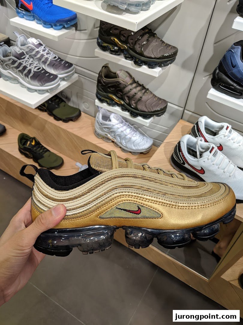 A hand holds a gold Nike sneaker in a JD Sports store, surrounded by other athletic shoes on display.