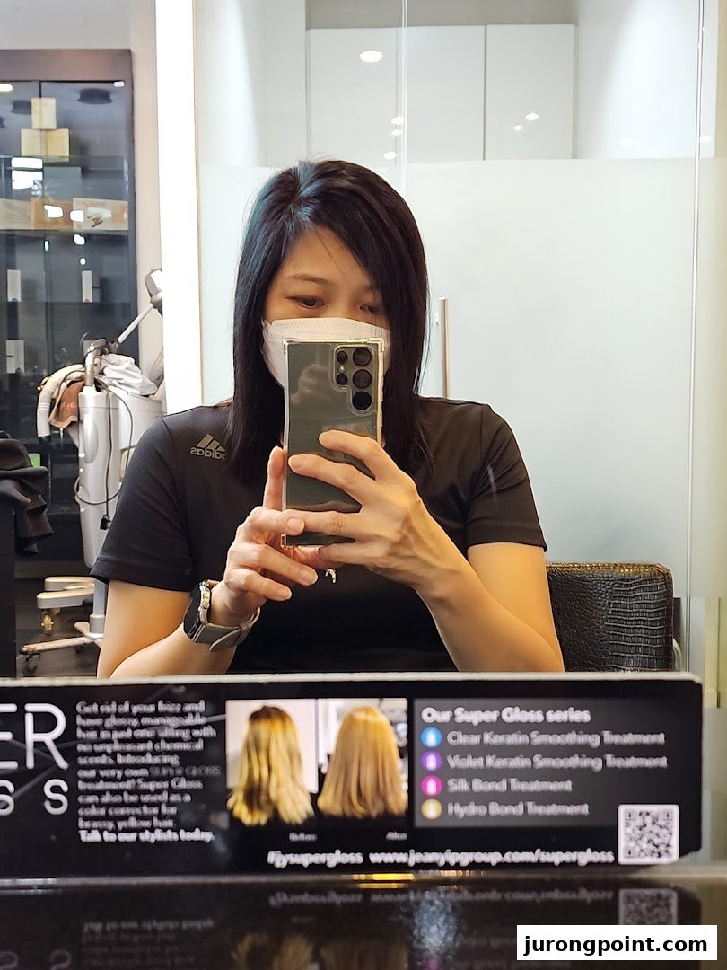A customer takes a selfie in a Jean Yip Aesthetics salon, featuring a display board for Super Gloss hair treatments.