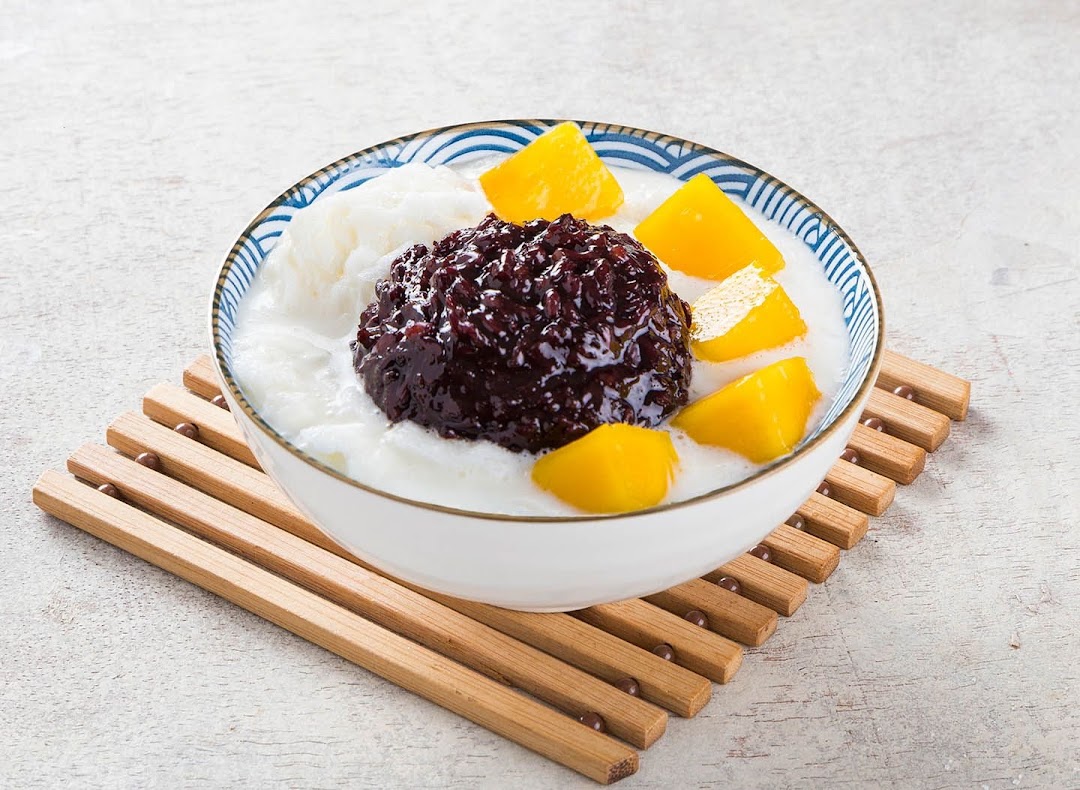 A vibrant bowl of dessert featuring black sticky rice, mango, and shaved ice on a wooden mat.