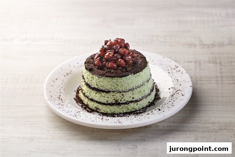 A delightful layered green dessert with chocolate crumbs and red bean topping on a white plate.