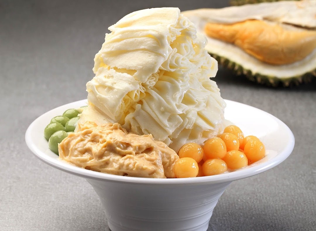 A bowl of creamy shaved ice dessert with green and orange toppings, and a durian in the background.