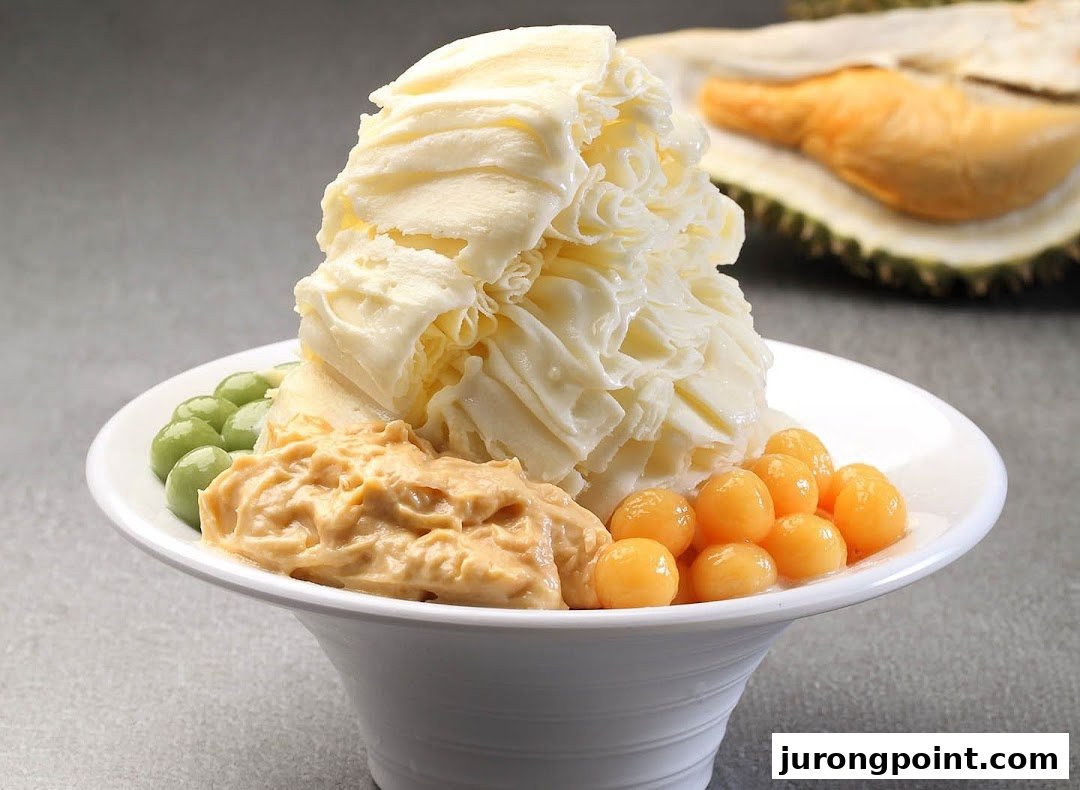 A bowl of creamy shaved ice dessert with green and orange toppings, and a durian in the background.