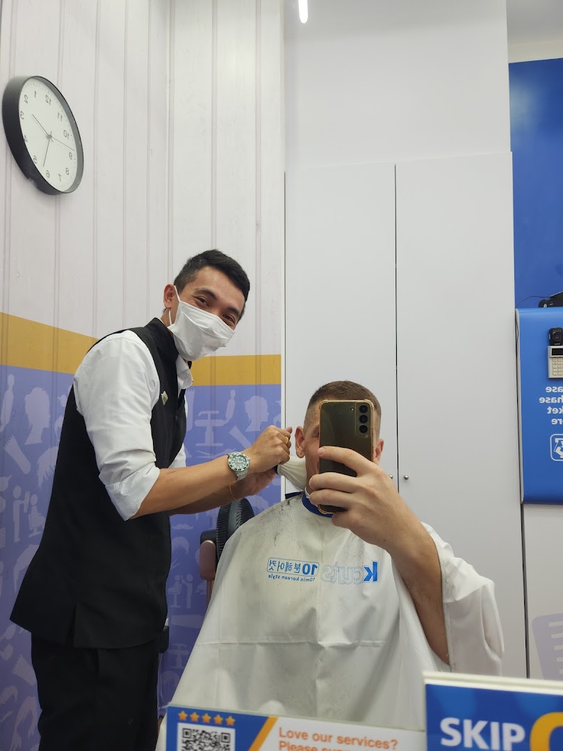 A barber wearing a mask cuts a customer's hair inside kcuts, while the customer takes a selfie.