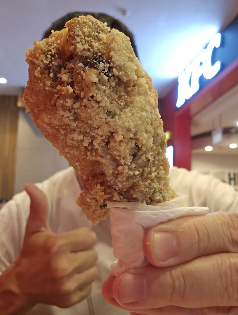 A hand holds a piece of crispy fried chicken, with a thumbs-up and KFC logo in the background.