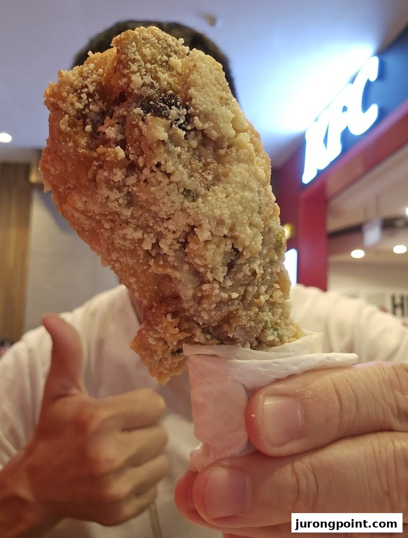 A hand holds a piece of crispy fried chicken, with a thumbs-up and KFC logo in the background.