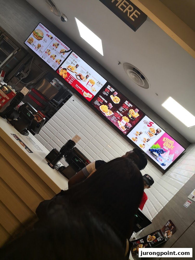 Digital menu boards display KFC's offerings above a busy counter with customers waiting to order.