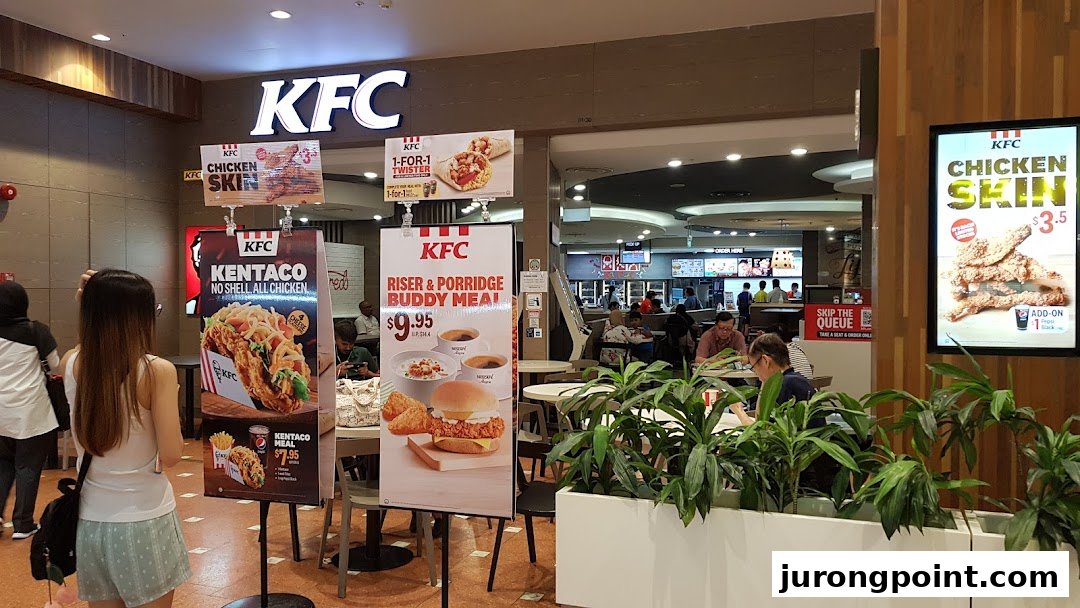 A bustling KFC outlet entrance in a mall, featuring promotional displays and customers dining inside.