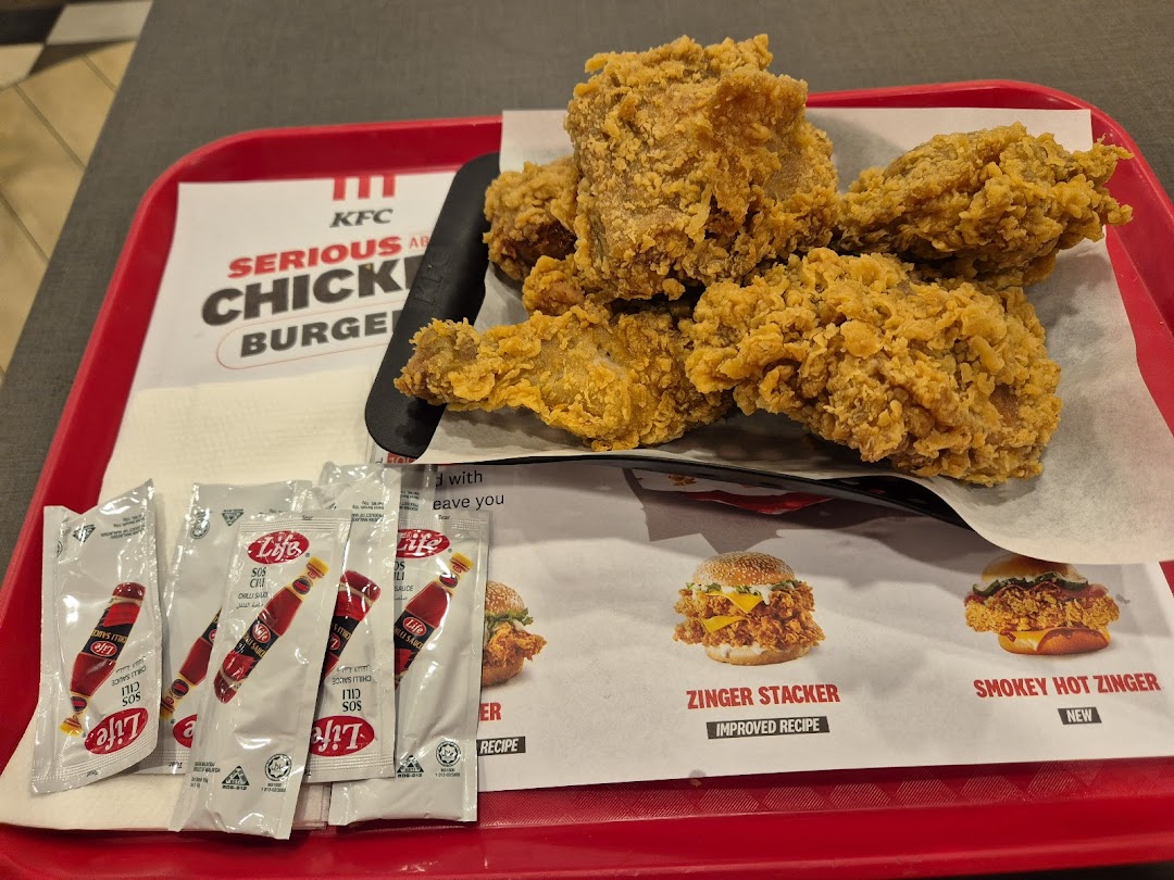 A tray of crispy KFC fried chicken with chili sauce packets and promotional material for Zinger burgers.