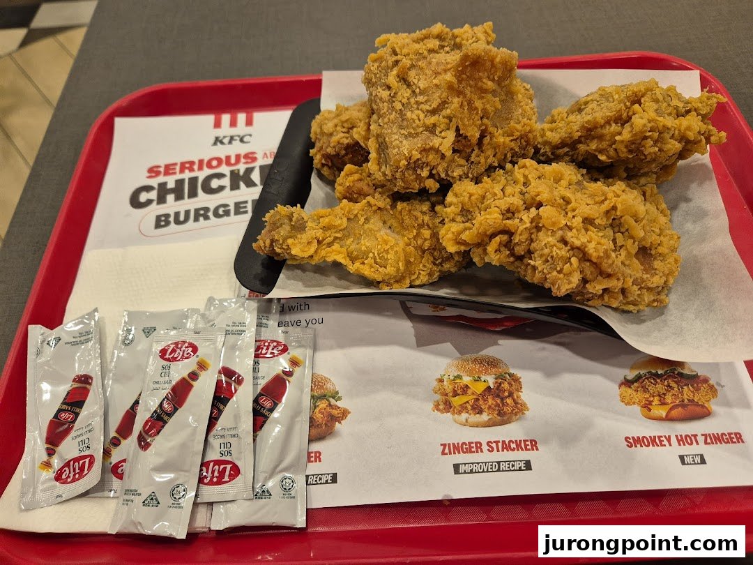 A tray of crispy KFC fried chicken with chili sauce packets and promotional material for Zinger burgers.