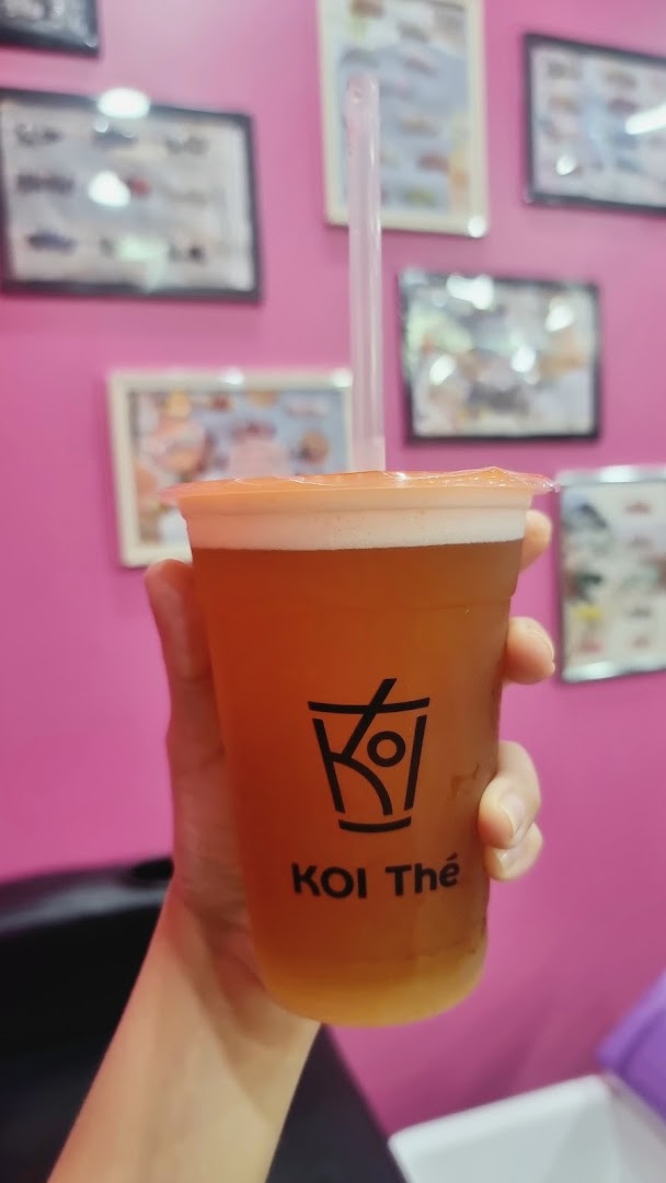 A hand holds a refreshing KOI Thé drink with a pink background and framed pictures.