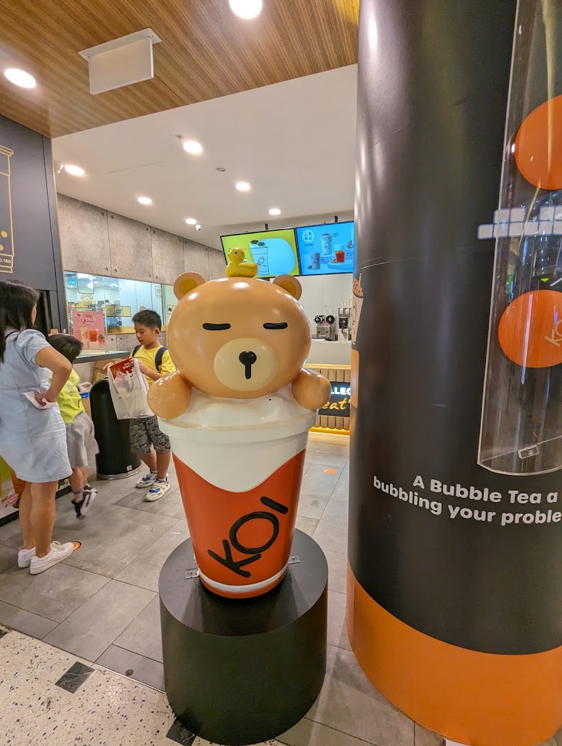 A large, cute bear mascot in a bubble tea cup stands at the entrance of a bustling KOI Thé shop.