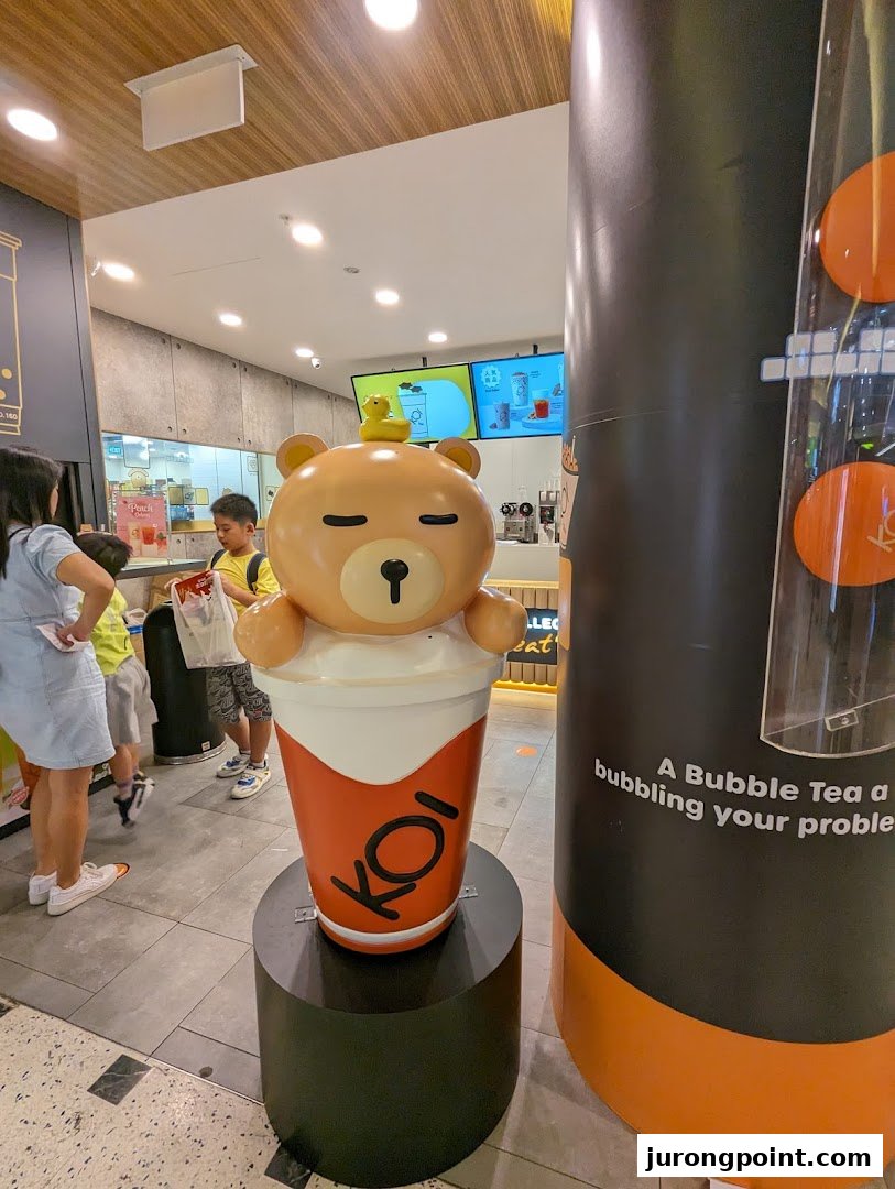 A large, cute bear mascot in a bubble tea cup stands at the entrance of a bustling KOI Thé shop.