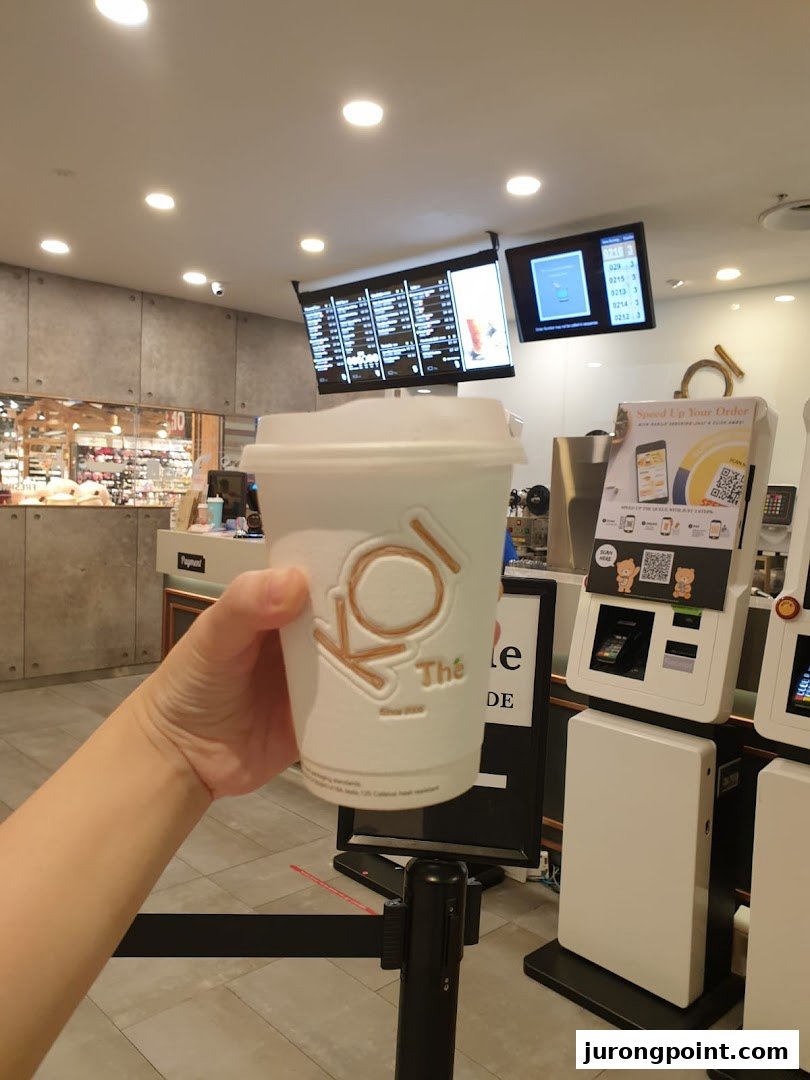 A hand holds a KOI Thé bubble tea cup inside the shop, with digital menus and self-service kiosks visible.