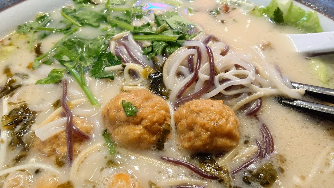 A close-up of a creamy noodle soup with meatballs, various noodles, and fresh greens.