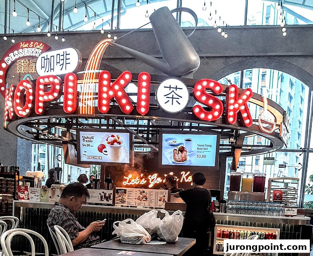 A vibrant Kopikiosk stall in a food hall, featuring neon signs, digital menus, and customers.
