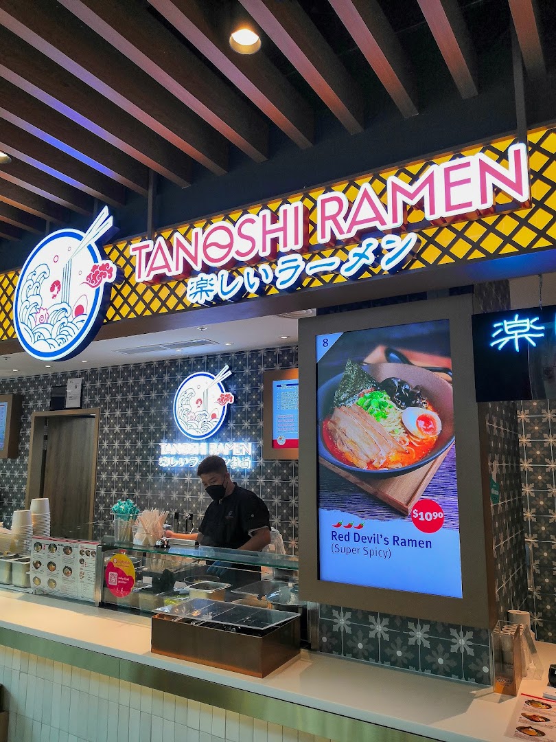 A vibrant counter at Tanoshi Ramen, featuring a staff member and a digital display showcasing their fiery Red Devil's Ramen.