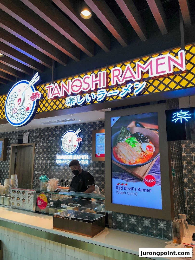 A vibrant counter at Tanoshi Ramen, featuring a staff member and a digital display showcasing their fiery Red Devil's Ramen.