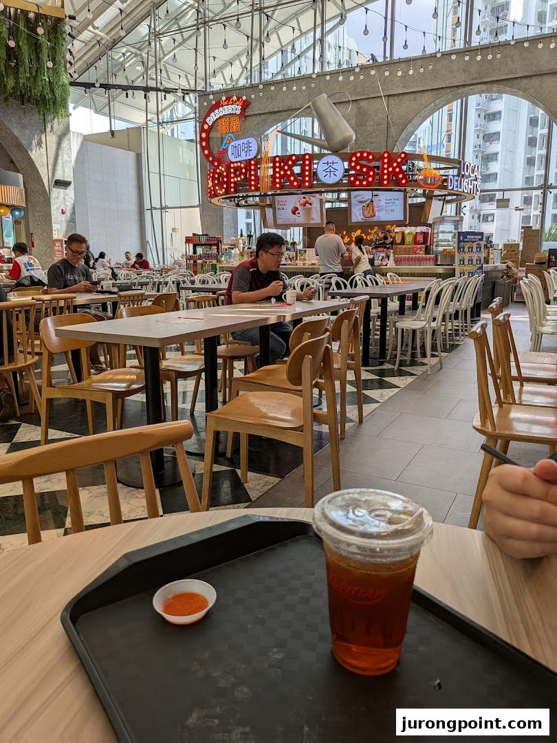 A vibrant interior view of Kopitiam Food Hall, featuring diners, tables, and a refreshing drink in the foreground.