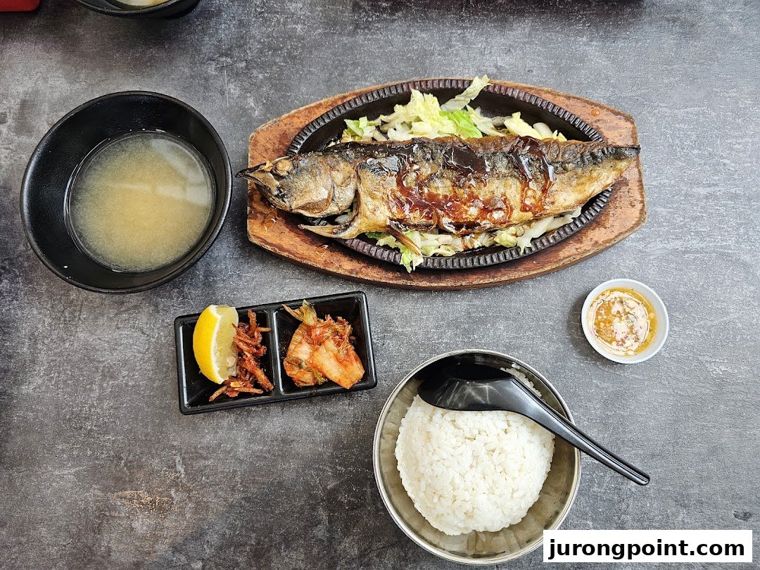A delicious grilled fish meal with rice, soup, and side dishes on a dark table.