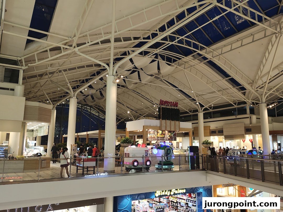 A spacious food hall with various dining options under a high, vaulted ceiling, featuring the Kopitiam sign.