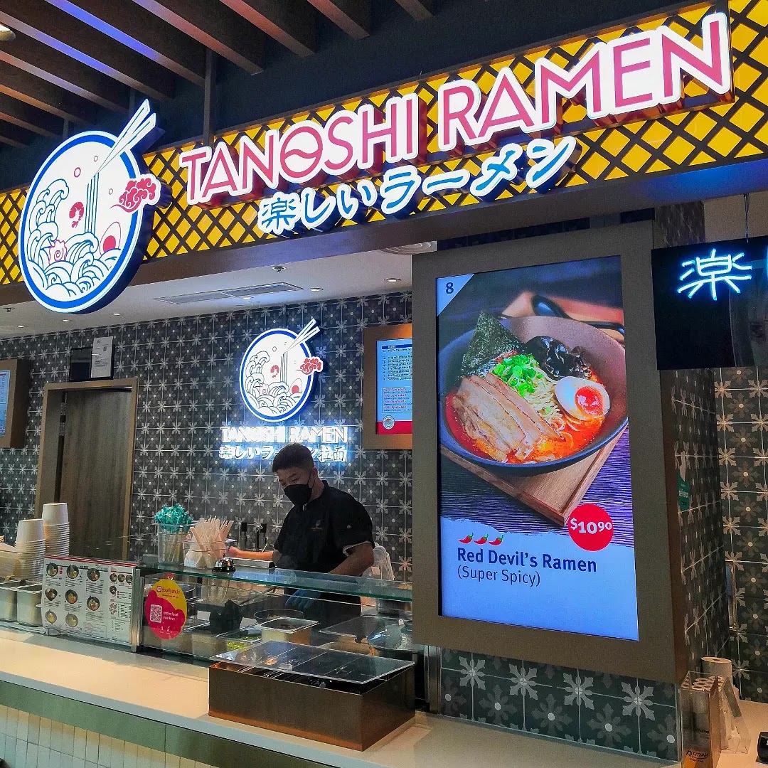 A vibrant ramen shop counter with a chef, prominent signage, and a digital display showcasing a spicy ramen dish.