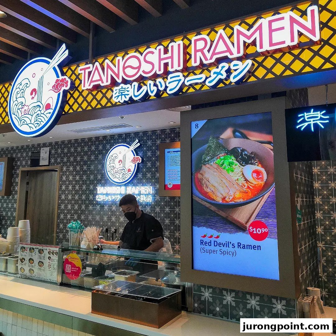 A vibrant ramen shop counter with a chef, prominent signage, and a digital display showcasing a spicy ramen dish.