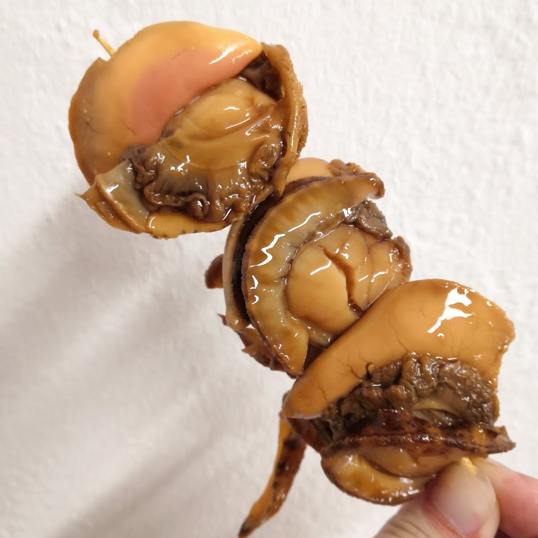 Close-up of glossy, cooked shellfish on a skewer, held by a hand against a white background.