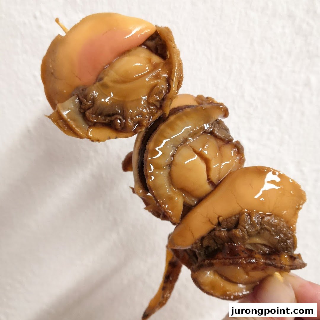 Close-up of glossy, cooked shellfish on a skewer, held by a hand against a white background.