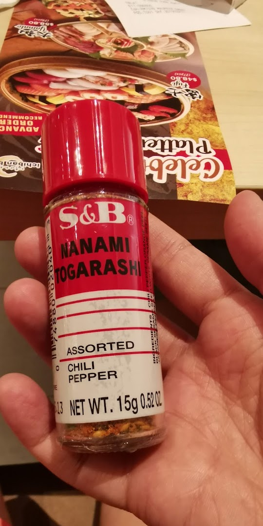 A hand holds a bottle of S&B Nanami Togarashi chili pepper, with a Japanese food platter menu visible in the background.