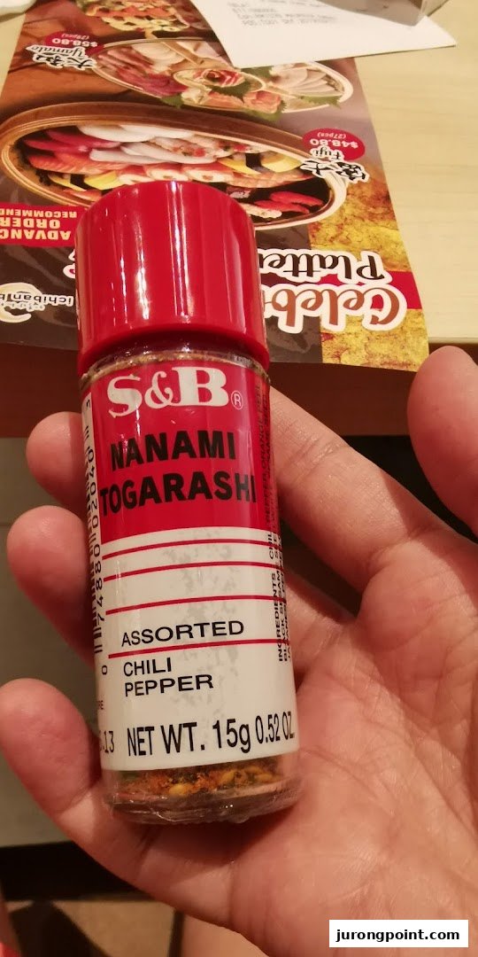 A hand holds a bottle of S&B Nanami Togarashi chili pepper, with a Japanese food platter menu visible in the background.