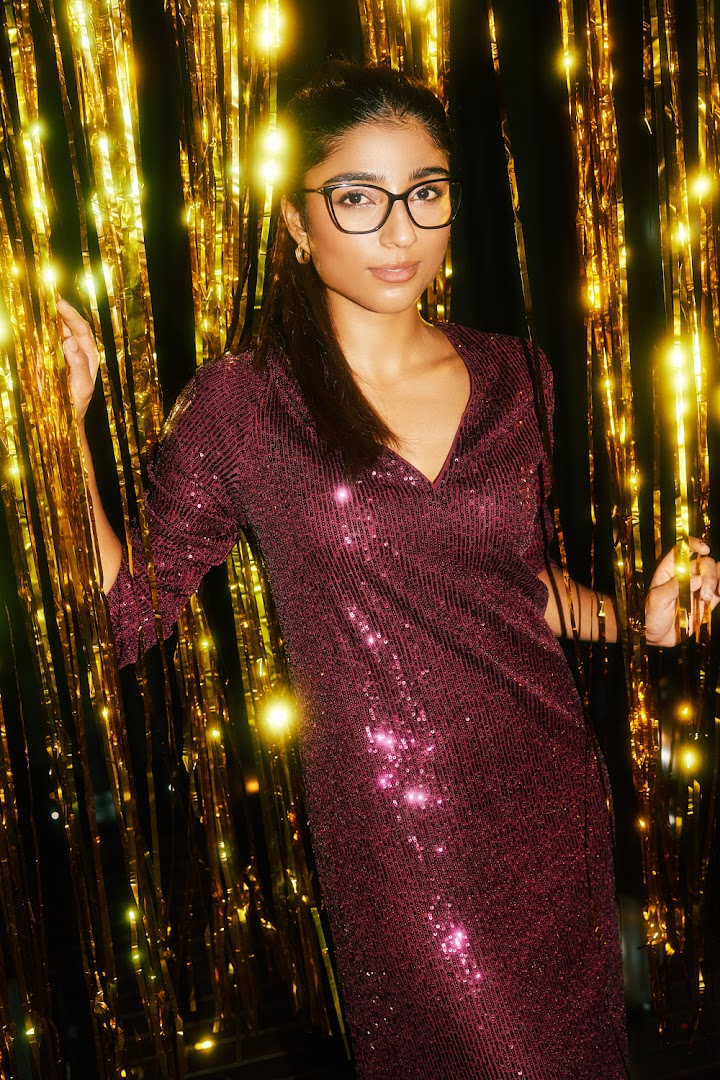 A woman wearing stylish black cat-eye glasses and a sequined dress against a golden backdrop.