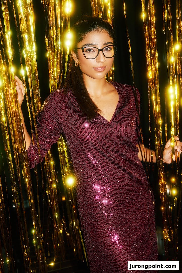 A woman wearing stylish black cat-eye glasses and a sequined dress against a golden backdrop.