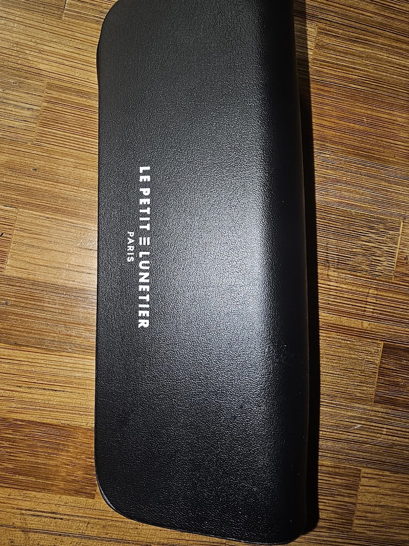 A black textured glasses case with 'LE PETIT LUNETTIER PARIS' branding on a wooden surface.