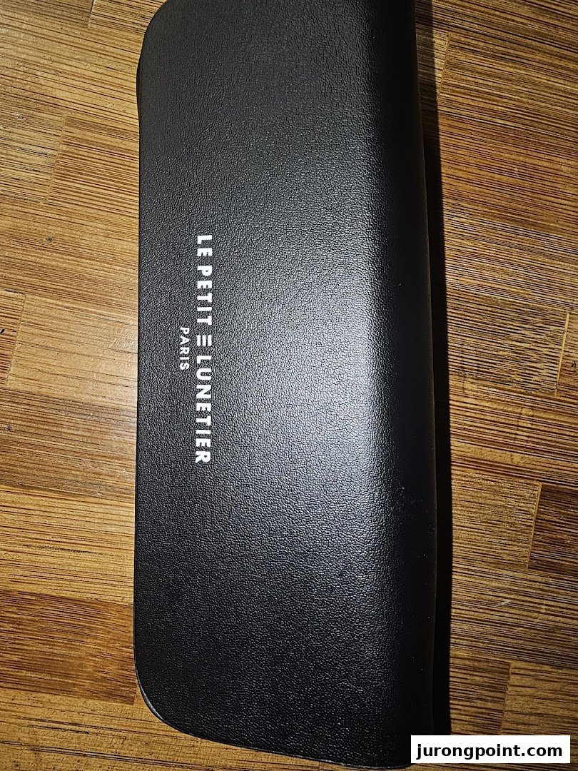 A black textured glasses case with 'LE PETIT LUNETTIER PARIS' branding on a wooden surface.