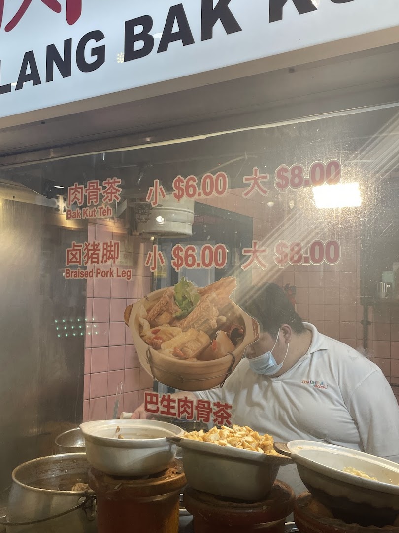A chef prepares dishes behind a glass counter displaying Bak Kut Teh and Braised Pork Leg prices.