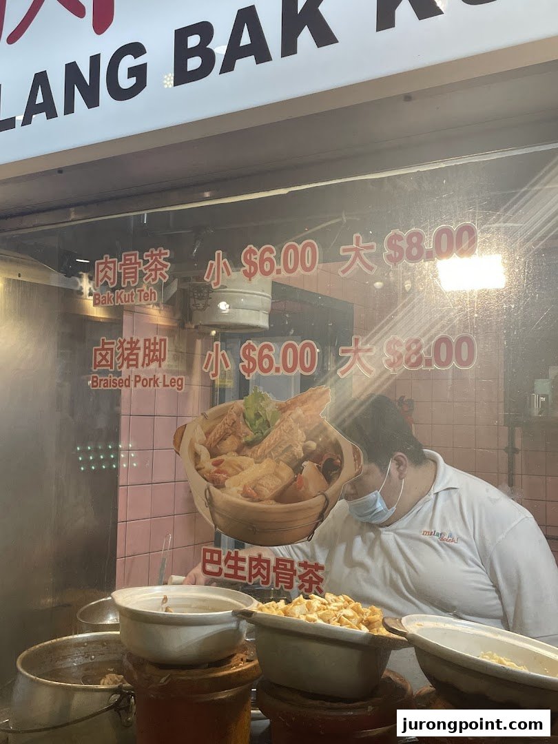 A chef prepares dishes behind a glass counter displaying Bak Kut Teh and Braised Pork Leg prices.