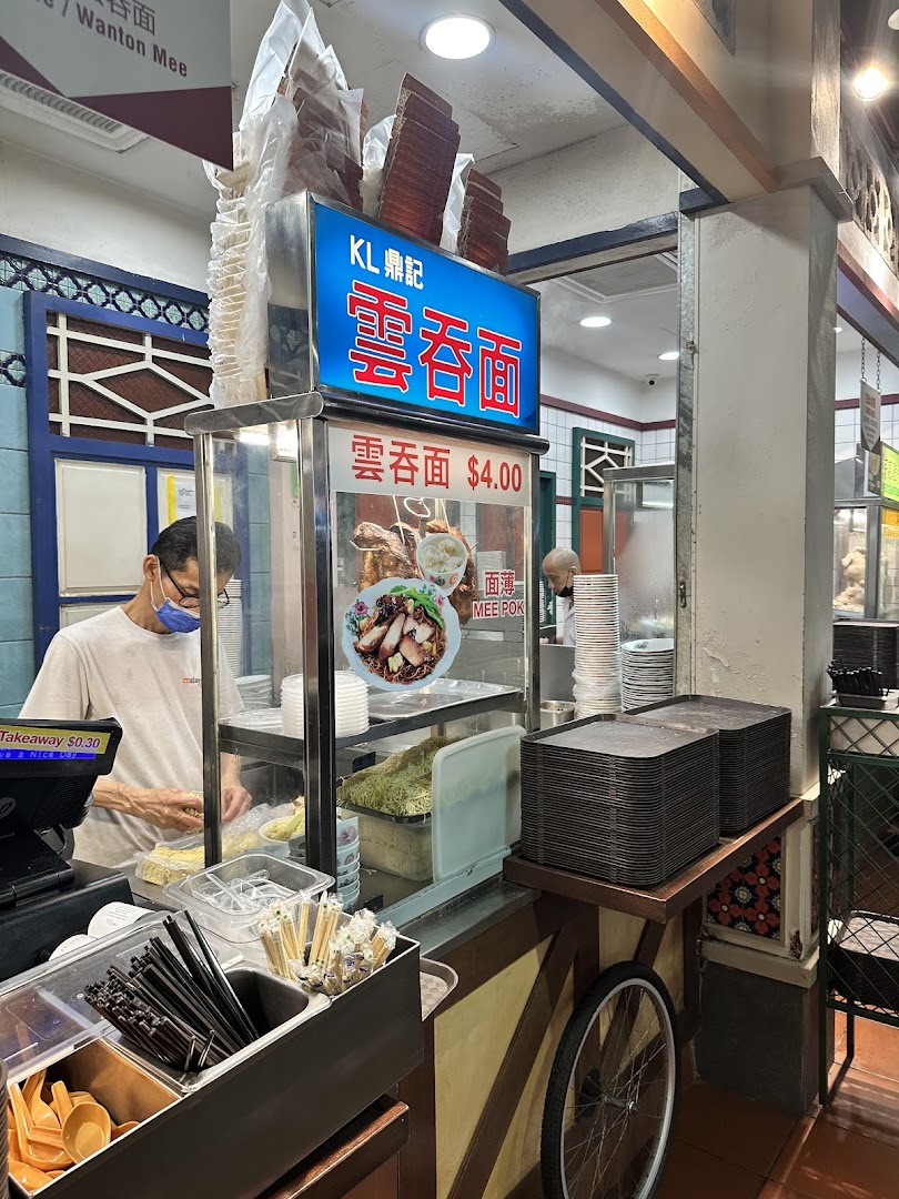 A bustling food stall counter with a chef preparing noodles, featuring signage for 'Wanton Mee' and 'Mee Pok'.