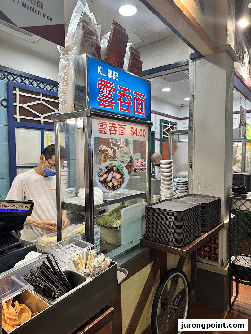 A bustling food stall counter with a chef preparing noodles, featuring signage for 'Wanton Mee' and 'Mee Pok'.