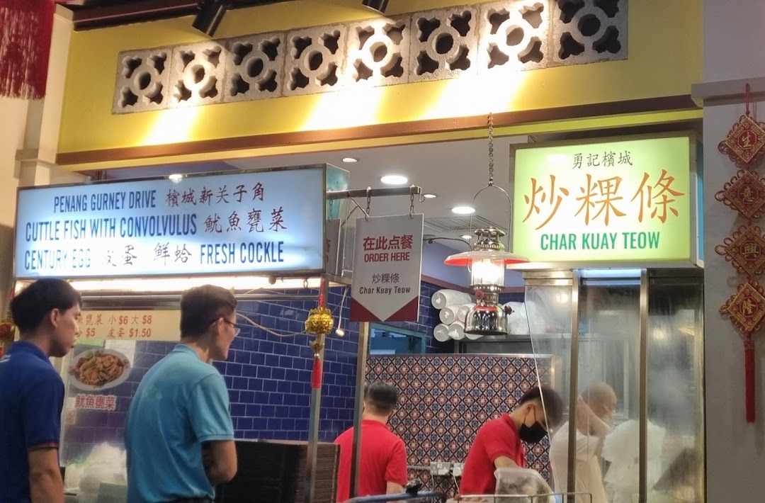 A bustling food stall counter featuring signs for Char Kuay Teow and other Malaysian dishes.