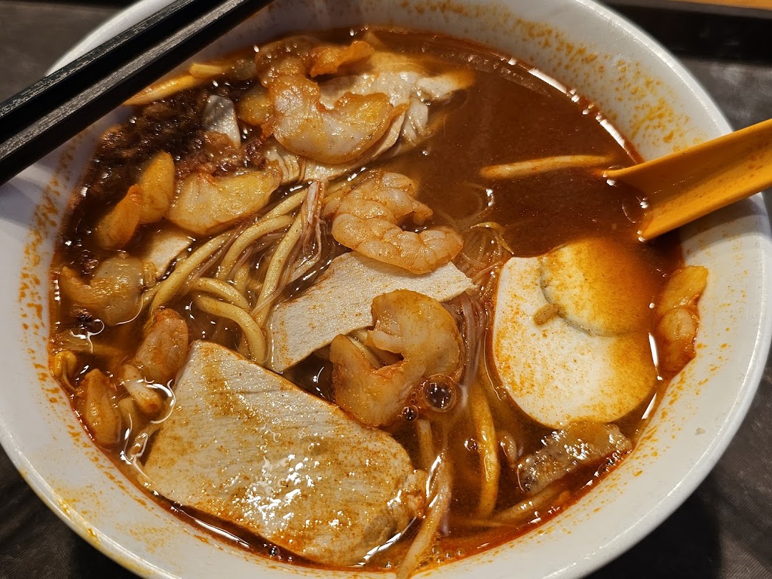 A close-up of a vibrant bowl of spicy Malaysian noodles with prawns, fish cake, and a rich broth.