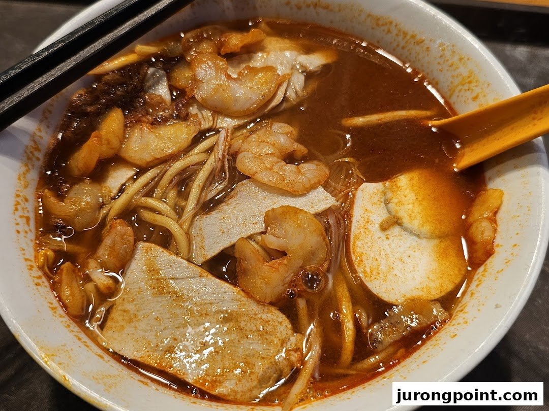 A close-up of a vibrant bowl of spicy Malaysian noodles with prawns, fish cake, and a rich broth.