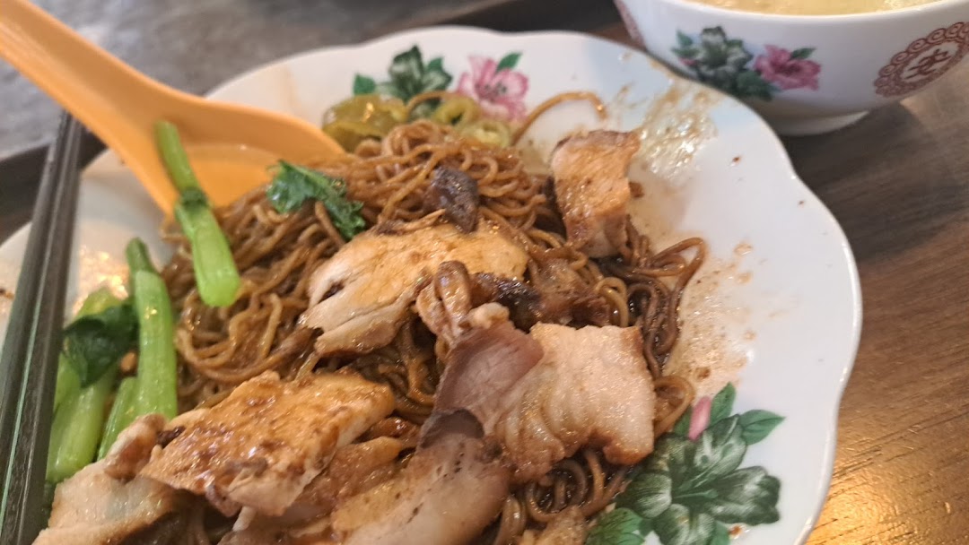 A close-up of delicious Malaysian-style noodles with roasted pork and greens on a floral plate.