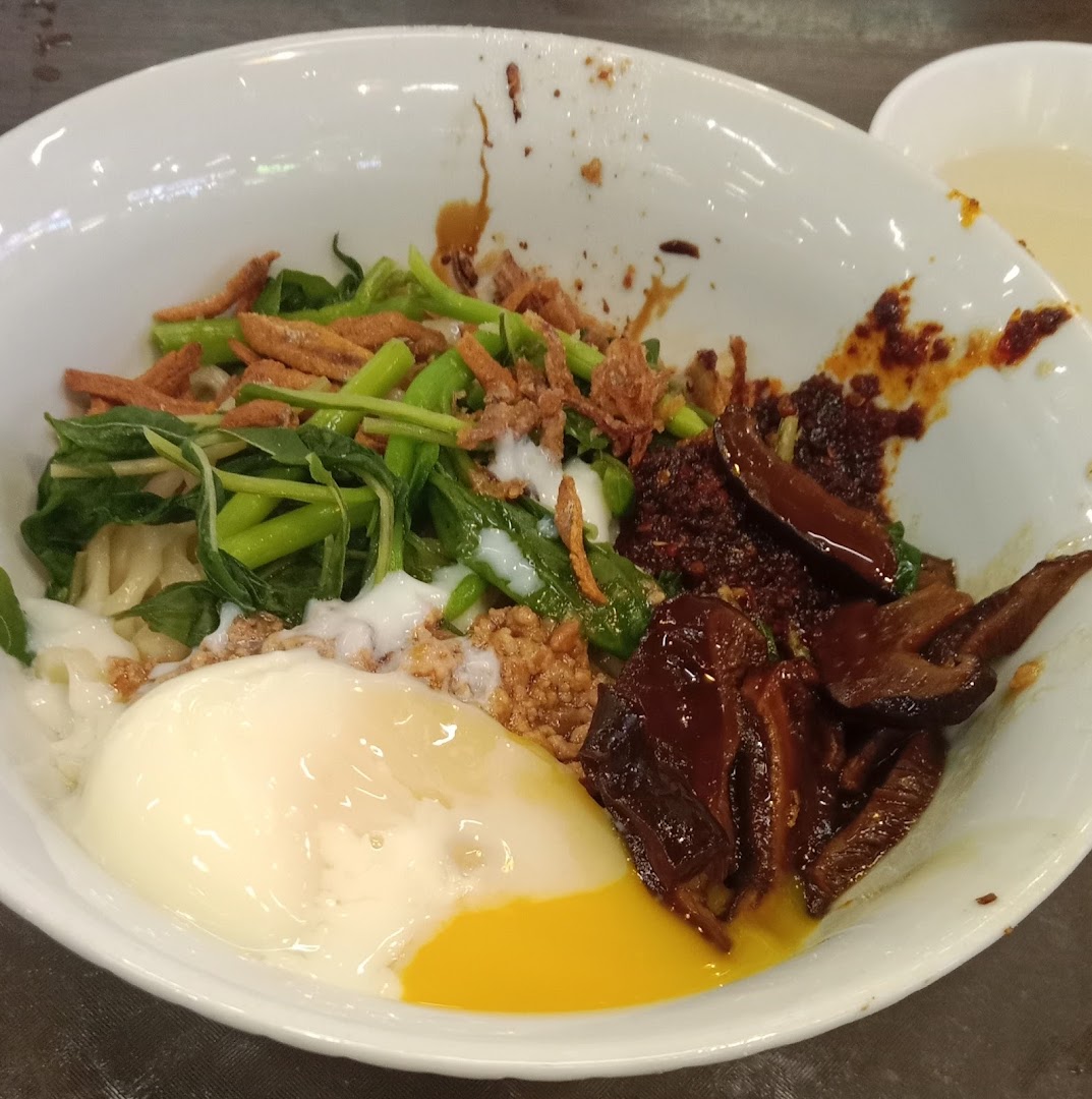 A vibrant bowl of Malaysian noodles with a runny egg, greens, mushrooms, and chili.
