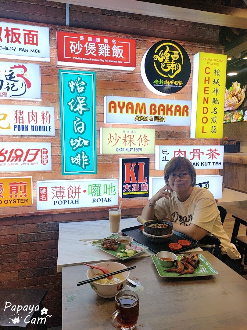 A person enjoying a variety of Malaysian dishes at a food court with numerous food stall signs.