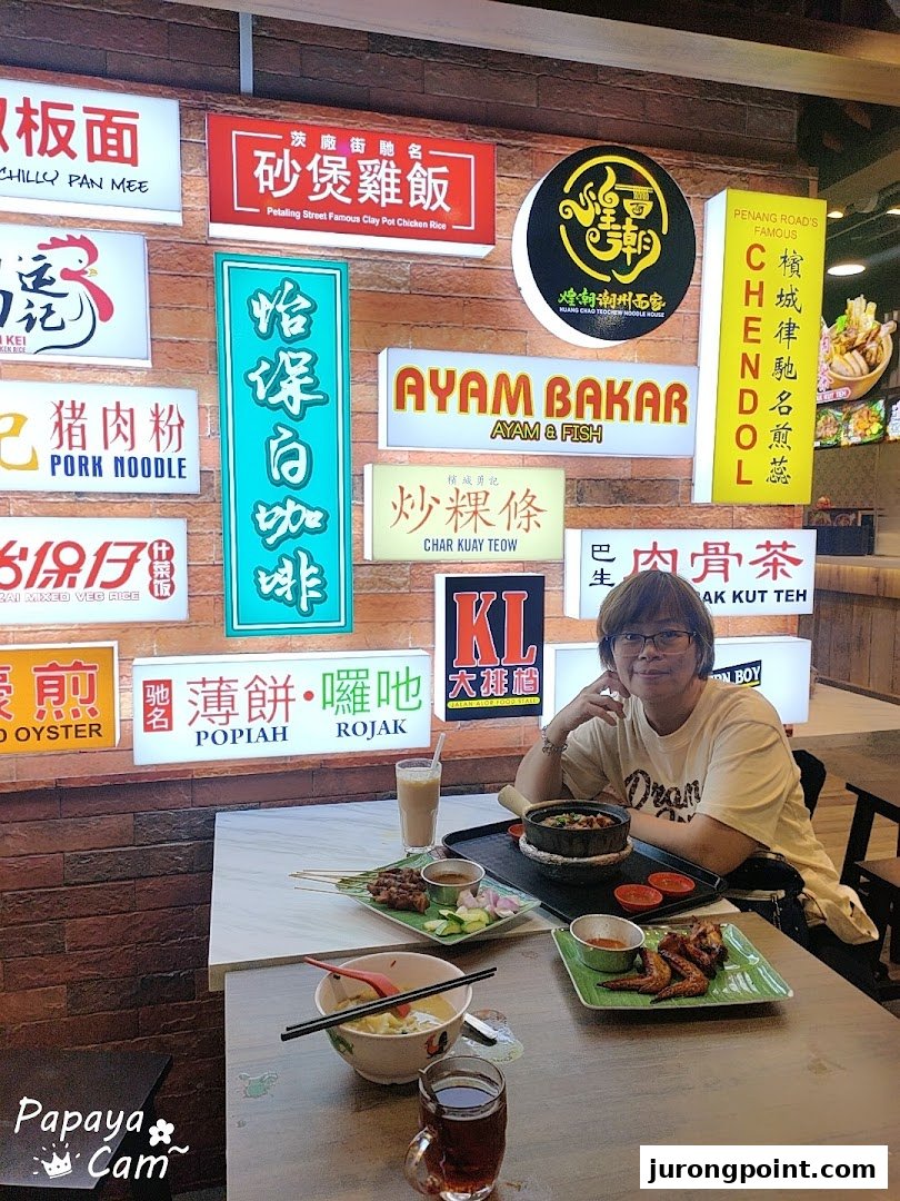A person enjoying a variety of Malaysian dishes at a food court with numerous food stall signs.